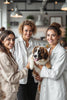 Three professional groomers smiling while holding a fluffy puppy in a modern grooming studio, representing grooming education, mentorship and early-career development.